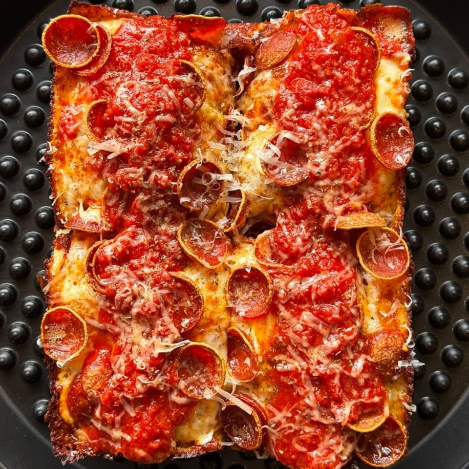 Square Detroit-style pizza with pepperoni, rich red sauce, and melted cheese in a black pan.