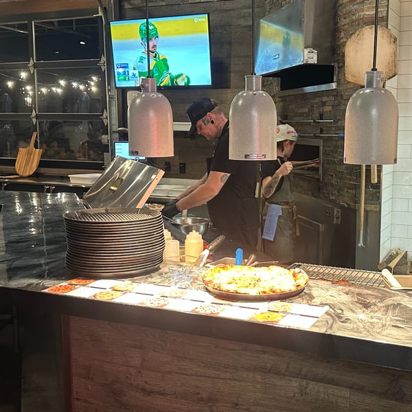 Serrano Brothers Eatery chefs preparing pizzas behind a counter with a finished pizza in the foreground.