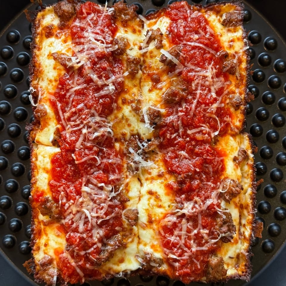 Square Detroit-style pizza with crispy crust, melted cheese, red sauce, and sausage crumbles.