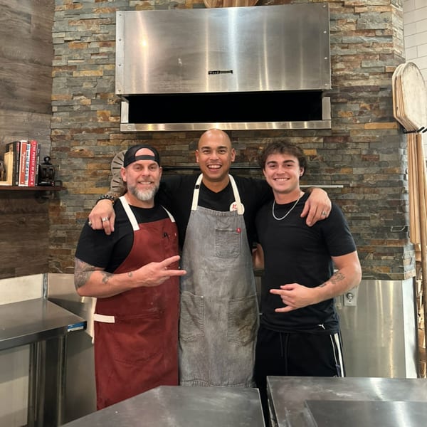 Three smiling Serrano Brothers Eatery chefs in aprons stand in front of a stone pizza oven.