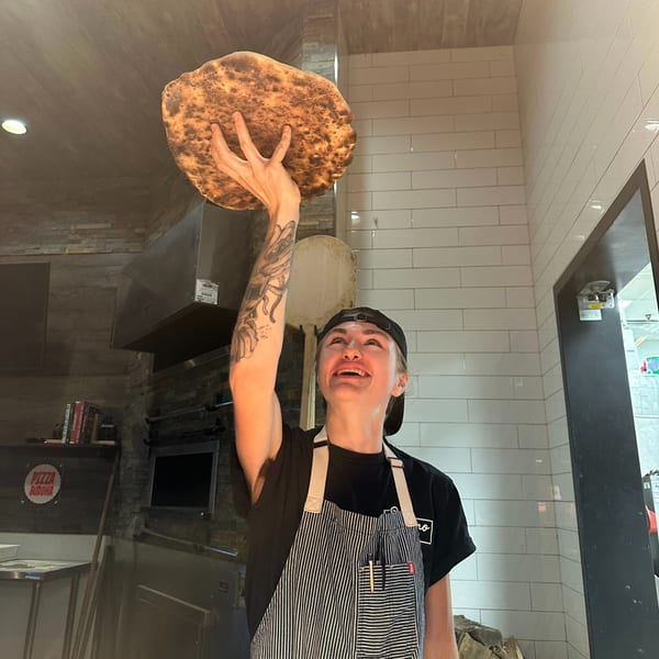 A smiling chef in an apron holds up a large, freshly baked pizza crust in a kitchen.